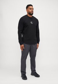 Zwarte gestructureerde sweatshirt met CK-logo, gecombineerd met donkergrijze broek en zwarte sportschoenen op een lichtgrijze achtergrond.