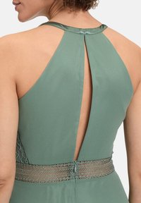 Zielona sukienka na halter z wycięciem w kształcie klucza na plecach oraz teksturowanym koronkowym pasem w talii. Gładki materiał o delikatnym połysku.
