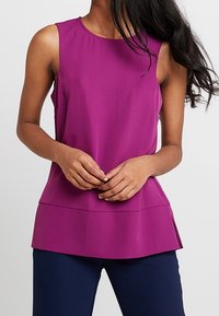 Fioletowa bluzka bez rękawów z gładkiego materiału z okrągłym dekoltem i warstwowym dołem. Zaprojektowana z luźnym krojem, z delikatnymi rozporkami po bokach.