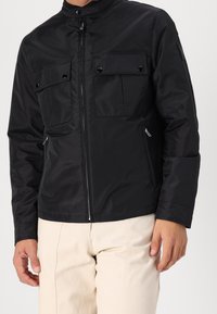 Černá nylonová bunda s stojacím límcem, dvěma náprsními kapsami na patentky, bočními kapsami na zip a zapínáním na zip vpředu.