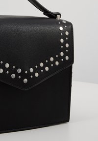Czarna skórzana torebka z srebrnymi ćwiekami na klapie, o wyraźnie uformowanym kształcie i gładkiej teksturze. Pasek w zestawie.