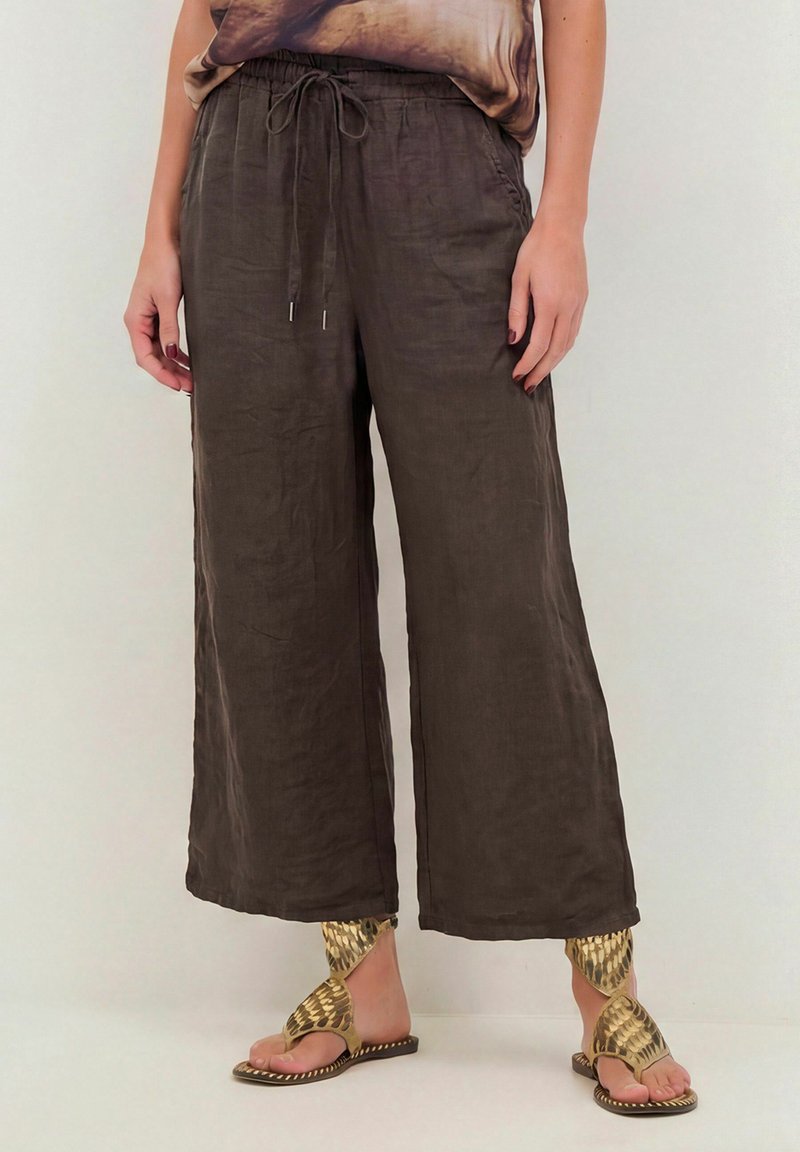 Femme portant un pantalon large en lin marron foncé avec une taille élastique et un cordon de serrage, associé à des sandales plates dorées texturées.