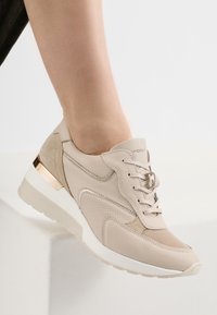 Scarpa sportiva beige realizzata in materiali in pelle e scamosciato, con design perforato, chiusura con lacci, dettagli metallici e una suola bianca spessa.