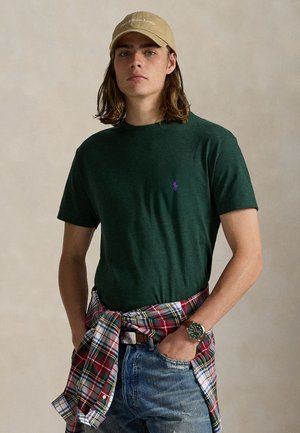 Joven con cabello largo que lleva una camiseta verde, gorra beige, vaqueros azules, reloj y una camisa de cuadros atada a la cintura, de pie con las manos en los bolsillos.