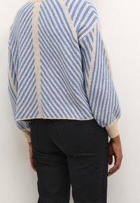 Pull en tricot beige et bleu à rayures, avec un design court, un col rond et des manches amples. Porté avec un pantalon noir.