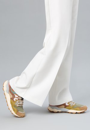 Legs in white wide-leg pants wearing multicolored suede and mesh sneakers with thick beige soles on a gray background.