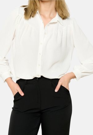 Kobieta ubrana w białą, zapinaną na guziki bluzkę z długim rękawem i bufkami na ramionach oraz czarne spodnie z wysokim stanem, z rękami w kieszeniach.