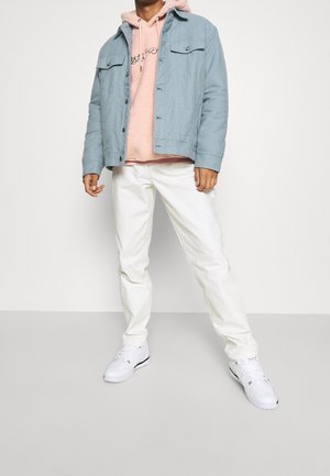 Giacca di jeans azzurro chiaro sopra una felpa con cappuccio rosa in pile, abbinata a pantaloni bianchi e sneakers bianche. Cuciture e tasche visibili.