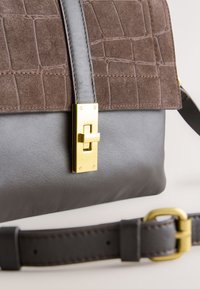 Sac à main avec fond en cuir gris foncé, dessus en daim marron avec texture crocodile, fermoir rectangulaire doré à tournant, et bandoulière amovible avec boucle.