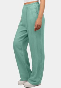 Lichtgroene, gestructureerde wijde broek met een elastische tailleband en zijzakken, gecombineerd met witte sneakers op een neutrale achtergrond.