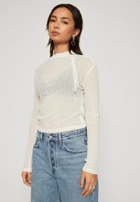 Bluză albă cu mâneci lungi, confecționată dintr-un material transparent și texturat, având un guler înalt și detalii cu șnur, asortată cu blugi din denim cu talie înaltă.