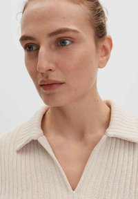 Helles beige gestricktes Oberteil mit Kragen und gerippter Textur, das subtile vertikale Muster und eine lockere Passform aufweist.