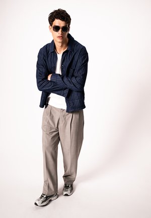 Ung mand iført mørkeblå jakke, hvid skjorte, beige bukser, grå gymnastik-sko og solbriller, stående med armene krydset mod en hvid baggrund.