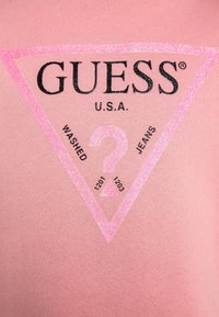 Camiseta de algodón rosa con un logo triangular en rosa brillante y negro con el texto "GUESS U.S.A." y los números 1201, 1203, "WASHED" y "JEANS."
