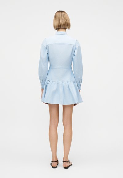 Γυναίκα που στέκεται πλάτη, φορώντας ένα ανοιχτό γαλάζιο μίνι φόρεμα με μακριά μανίκια και φουσκωτή φούστα, καθώς και μαύρα σανδάλια με επίπεδη σόλα.