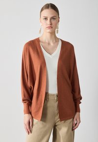 Rustfarvet strikket cardigan med åben front, afslappet pasform og lange ærmer, lagd oven på en hvid v-hals top og beige bukser.