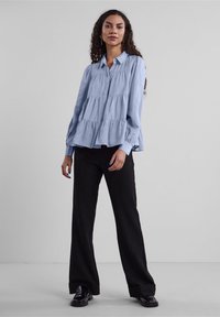 Hellbluse mit gestuften Rüschendesign, Knopfkragen, langen, geknöpften Ärmeln, getragen mit schwarzen, weiten Hosen und schwarzen Schuhen.