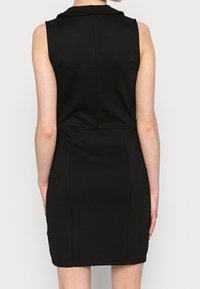 Robe noire sans manches avec un col, taille cintrée et ourlet droit, vue de dos sur une personne se tenant devant un fond uni.