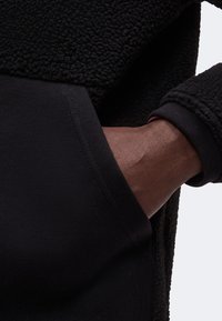 Main à la peau foncée partiellement insérée dans la poche de la manche d’une veste polaire noire texturée.