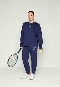 Tumši zils džemperis un atbilstoši sporta bikses no mīkstas auduma, ar gaiši zilu logo. Rokā tur tennis raketi un bumbu, ar baltām kedām.