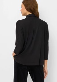 Frau mit braunen Haaren, die ein lockeres schwarzes Oberteil mit Dreiviertelärmeln und schwarze Hosen trägt, seitlich vor einem weißen Hintergrund.