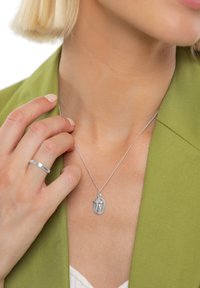 Sidabrinė pakabuko grandinėlė su sudėtingu religinio dizaino motyvu, kabančiu ant plonos grandinėlės. Nešiojama ant žalsvos švarkelio, ranka švelniai liečia pakabutį.