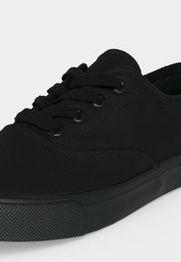 Nahaufnahme eines schwarzen Low-Top-Sneakers mit schwarzen Schnürsenkeln, Metallösen und einer strukturierten Gummisohle und Zehenkappe.