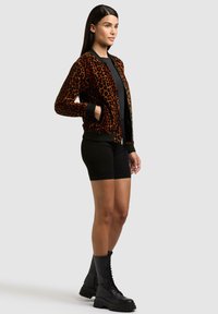 Femme debout portant une veste à imprimé léopard, un short noir et des bottes de combat noires lacées, sur un fond gris clair uni.