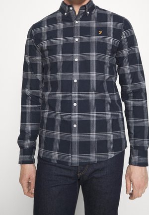 Mann trägt ein marineblaues, langärmeliges Hemd mit weiß kariertem Muster und einem kleinen gelben, gestickten Logo auf der Brust.