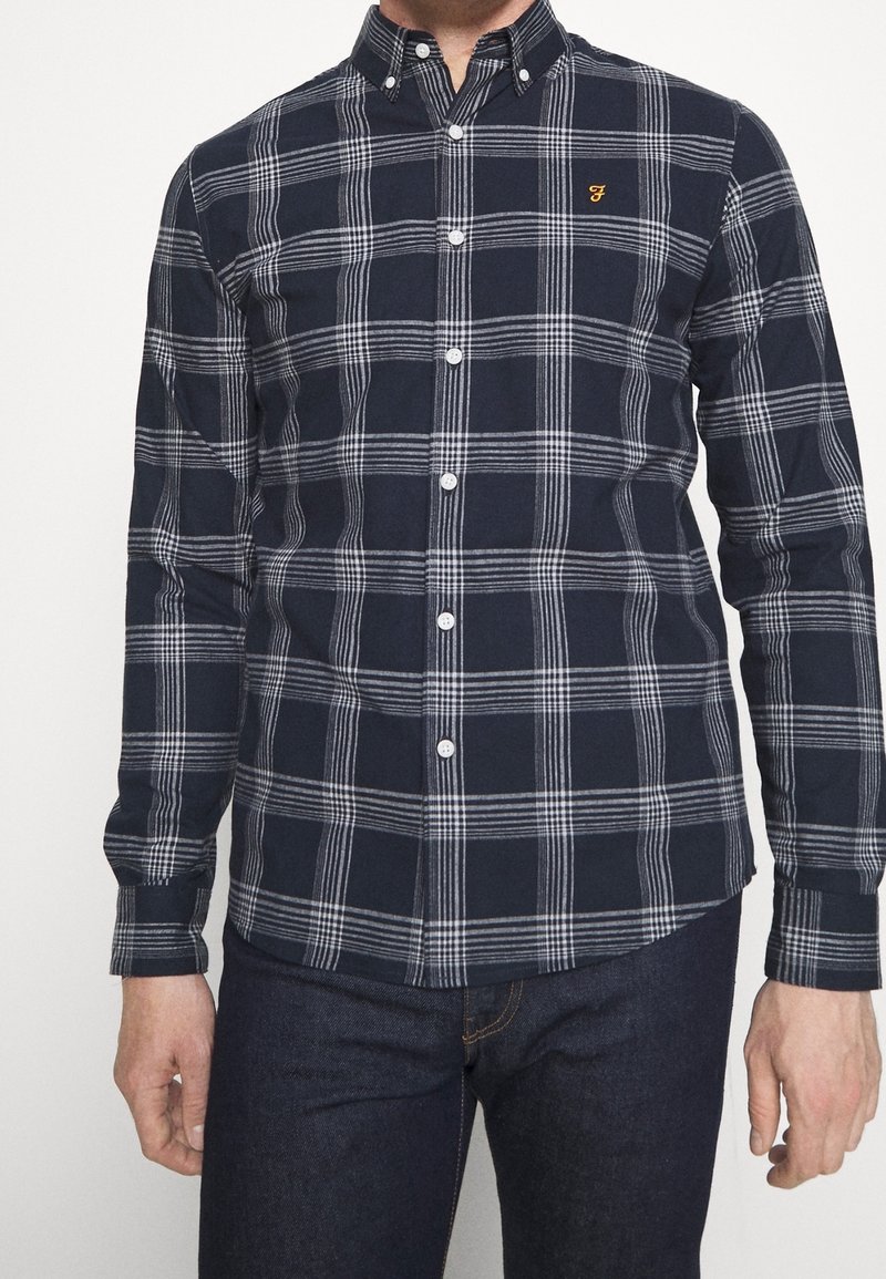 Homme portant une chemise boutonnée à manches longues bleu marine avec un motif à carreaux blancs et un petit logo brodé jaune sur la poitrine.