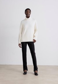 Weißer, gerippter Rollkragenpullover kombiniert mit schwarzen, maßgeschneiderten Hosen und schwarzen, hochhackigen Schuhen. Glatte Textur und lockere Passform mit überschnittenen Schultern.