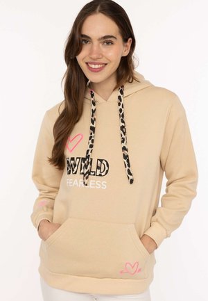 Junge Frau lächelt, trägt einen beigen Hoodie mit Leopardenmuster-Kordeln und dem Schriftzug „WILD FEARLESS“ mit pinkfarbenen Herzmotiven.
