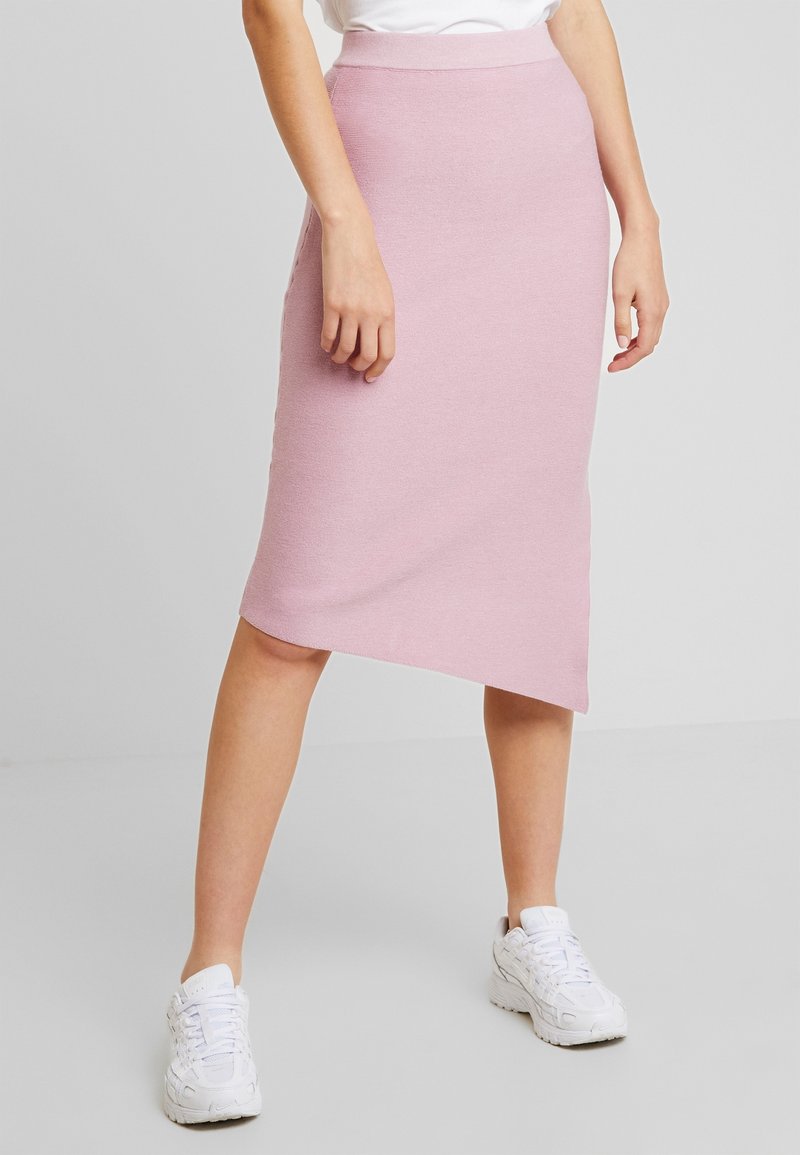 Pletená midi sukně v jemně růžové barvě s asymetrickým lemem, vybavená hladkou texturou a přiléhavým pasem. Nošeno s bílými teniskami.