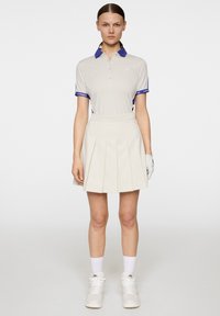 Ljust beige polo med blå krage och manschetter, kombinerat med en plisserad beige kjol. Vita sneakers och handskar fulländar outfiten.