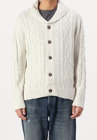 Krämfärgad stickad cardigan med sjalkrage, prydd med ett kabelstickat mönster, fem bruna knappar samt ribbade muddar och nederkant. Bärs över mörka jeans.