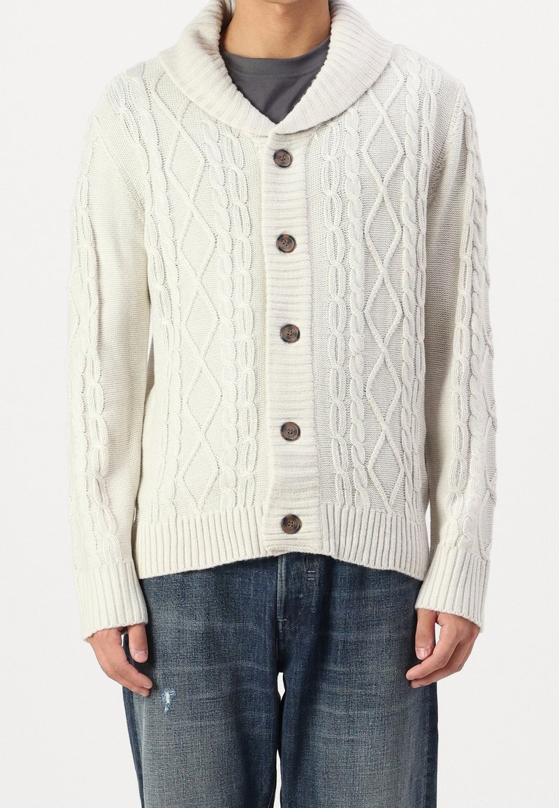 Krämfärgad stickad cardigan med sjalkrage, prydd med ett kabelstickat mönster, fem bruna knappar samt ribbade muddar och nederkant. Bärs över mörka jeans.