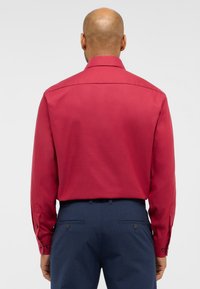 Camicia rossa a maniche lunghe con colletto, caratterizzata da una texture liscia. Abbinata a pantaloni blu scuro con tasche posteriori e dettagli con bottoni.