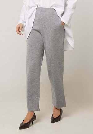 Vrouw draagt grijze, hooggetailleerde gebreide broek met zijzakken, gecombineerd met zwarte pumps met spitse neus en een losse witte blouse.