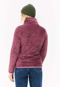 Chlpatá, tmavoružová fleecová bunda s vysokým golierom, doplnená zelenou pletenou čiapkou; nosená s modrými džínsami a svetlým topom pod ňou.