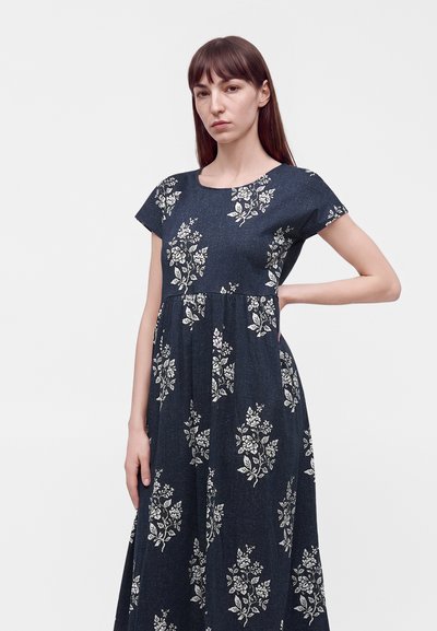 Femme aux longs cheveux bruns portant une robe bleu foncé à motifs floraux blancs, debout avec une main sur la hanche sur un fond uni.
