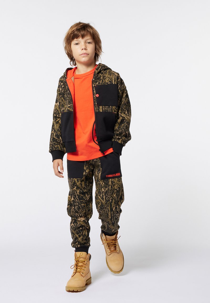 Felpa nera e in camouflage con tasche, maglietta arancione brillante, pantaloni in camouflage e stivali beige con lacci gialli.