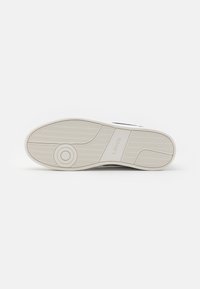 Biała podeszwa sportowego buta z teksturowanym wzorem, okrągłym logo i brandingiem na boku. Zaprojektowana z kontrastową granatową cholewką i trwałym materiałem.