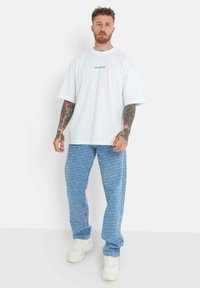 Camiseta blanca de gran tamaño con logo azul, combinada con pantalones de mezclilla azul claro que presentan un patrón de texto repetido. Zapatillas blancas completan el look.