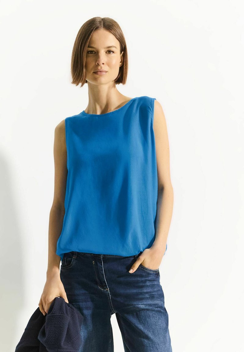 Top blu senza maniche in tessuto leggero con scollatura tonda. Abbinato a jeans di denim scuro e tiene un maglione blu navy. Design semplice.