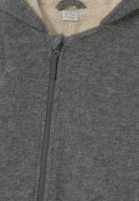 Tuta in felpa grigia con una morbida fodera interna beige, chiusura con zip frontale e cappuccio, caratterizzata da un'esterno testurizzato per calore e comfort.