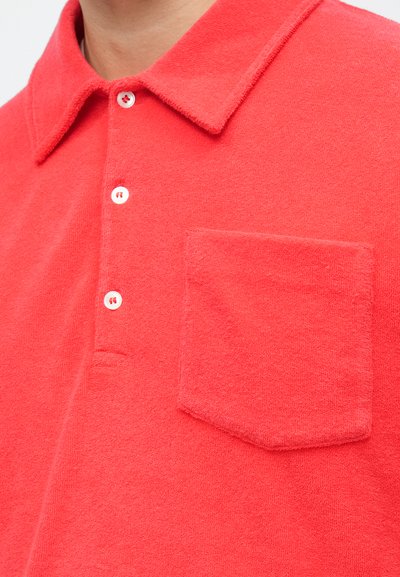 Polo rouge à manches courtes en tissu doux, avec un col pointu, trois boutons et une poche poitrine gauche.