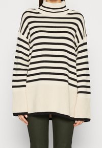 Sweter dżersejowy w paski z wysokim dekoltem w kolorze czarnym i kremowym. Charakteryzuje się luźnym krojem, szerokimi rękawami oraz żebrowanym wykończeniem na dole.