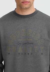 Sweatshirt gris avec un texte circulaire "QUALITÉ SUPÉRIEURE" en jaune et noir, avec les détails "DENIM FABRIQUÉ" et "Fournitures de biens secs". Tissu en mélange de coton.
