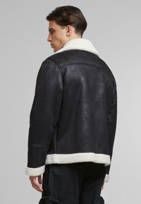 Chaqueta de shearling negra con cuello y dobladillo de piel sintética blanca. Presenta un corte relajado y un acabado texturizado, con mangas largas y sin herrajes visibles.