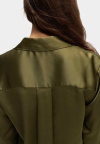 Olivengrüne Satinbluse mit glatter Textur, klassischem Kragen und gefälteltem Rückendetail, das das Licht mit einem glänzenden Finish einfängt.